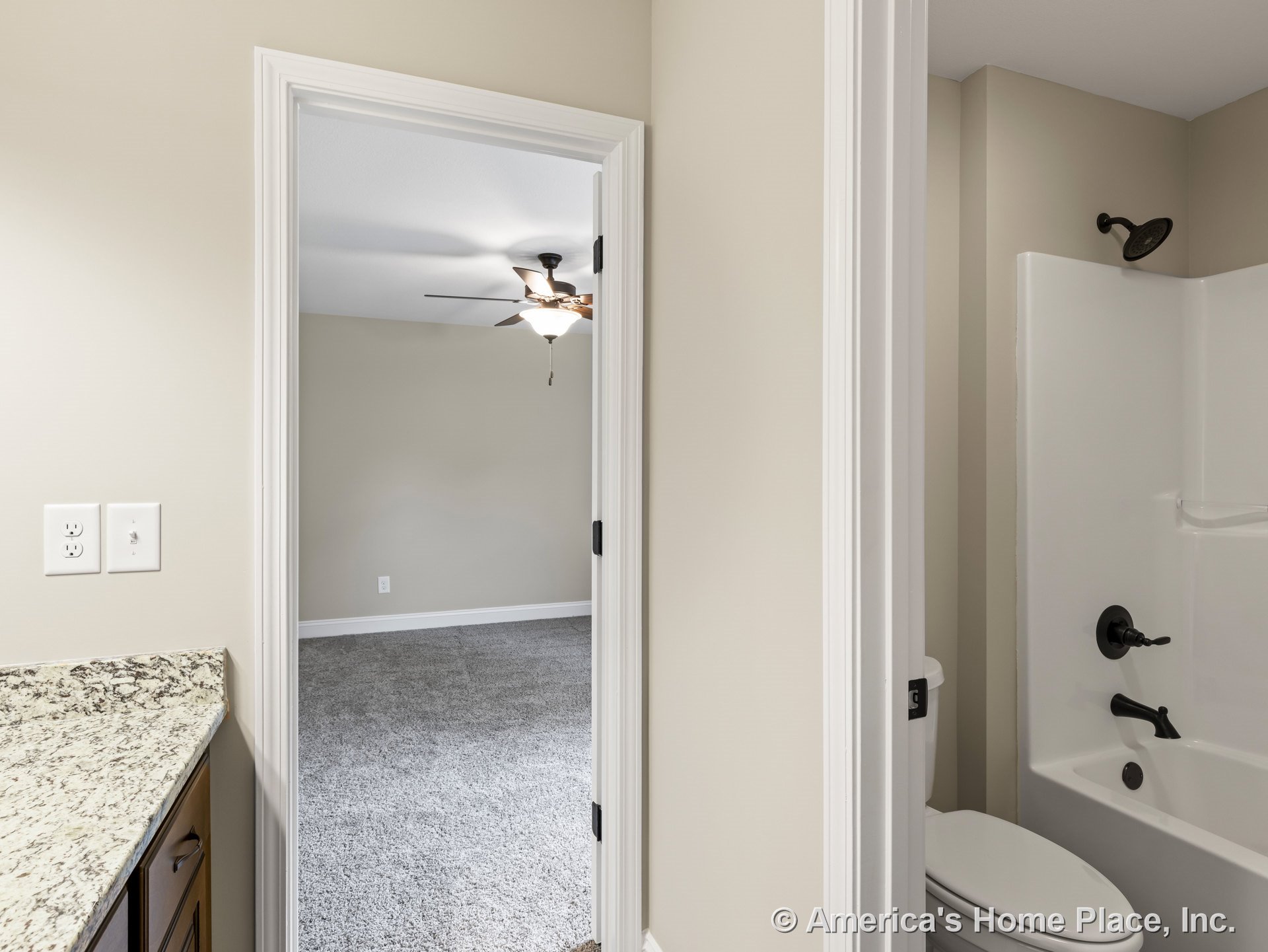 Granite bathroom countertop with dark wood cabinetry, white bathtub and shower, adjacent carpeted bedroom featuring ceiling fan with light fixture, neutral wall paint, and white