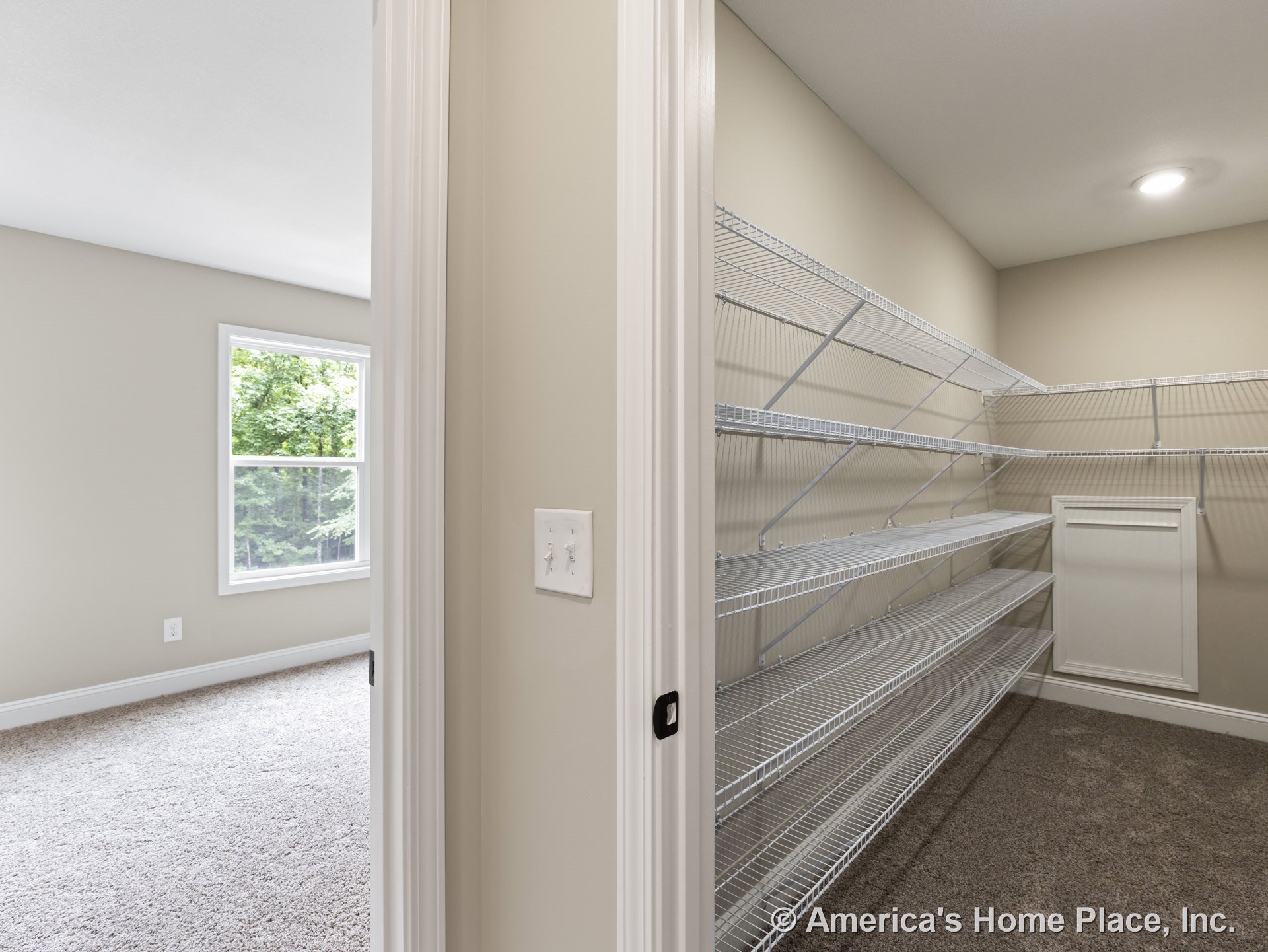 Walk-in closet featuring built-in wire shelving and recessed ceiling light, adjacent to carpeted bedroom with large window framed by white trim, neutral wall paint, and white