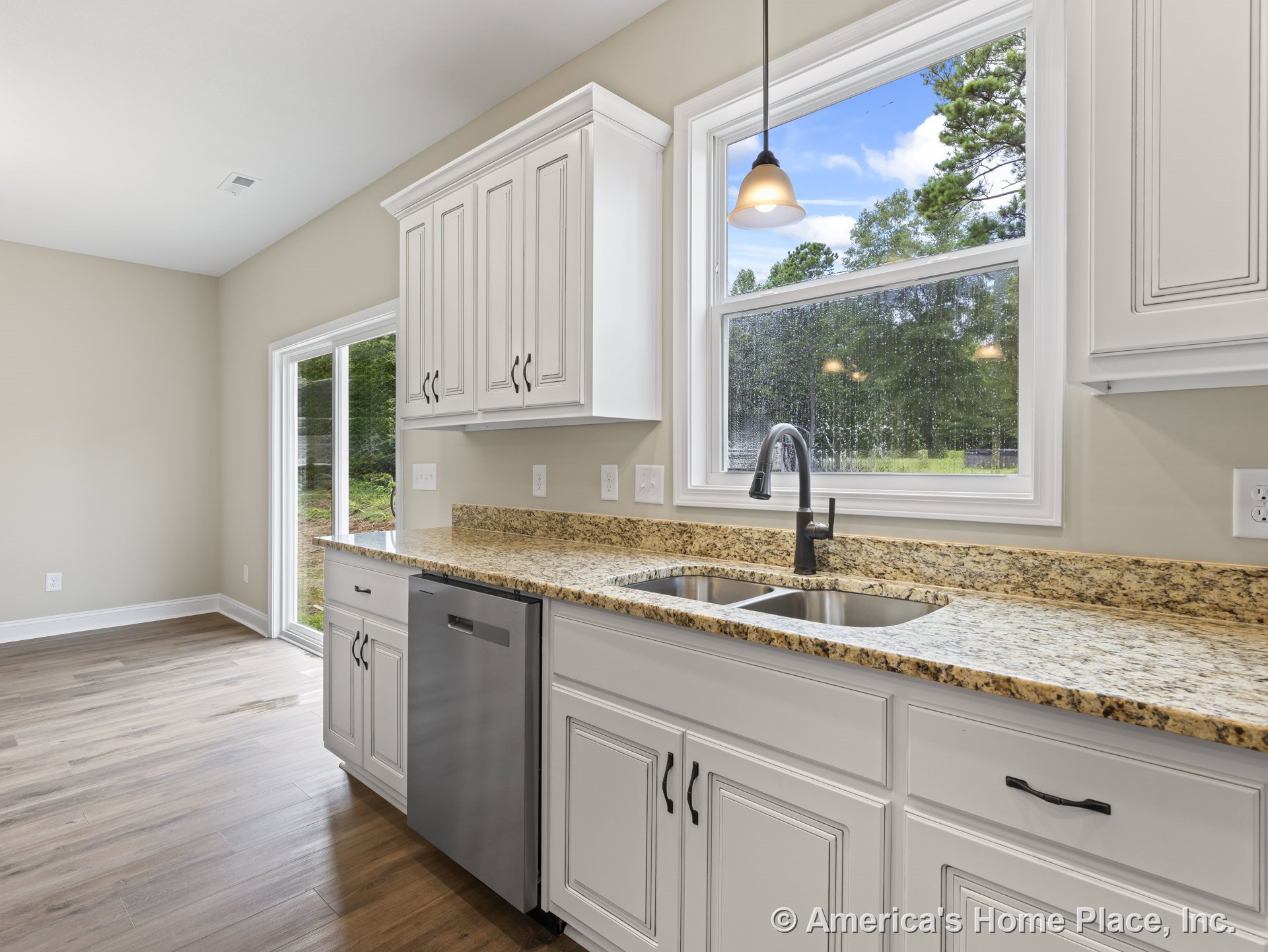 White upper and lower cabinets with granite countertops, double stainless steel sink set beneath a large window, pendant light fixture overhead, wood-look flooring, built-in
