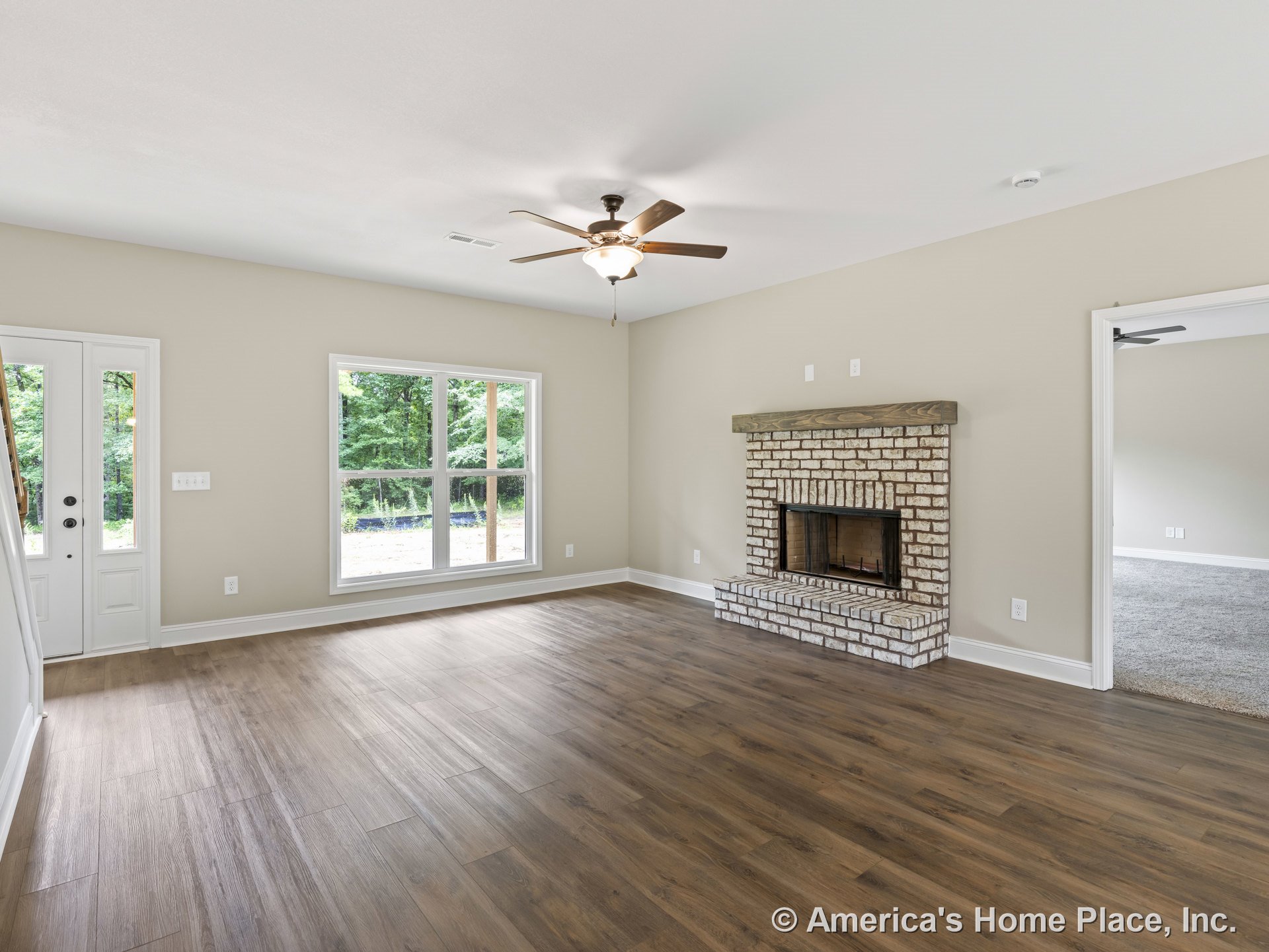 Brick fireplace with wood mantel, double window with white trim, wood-look plank flooring, ceiling fan with light fixture, neutral wall paint, and white double front doors.