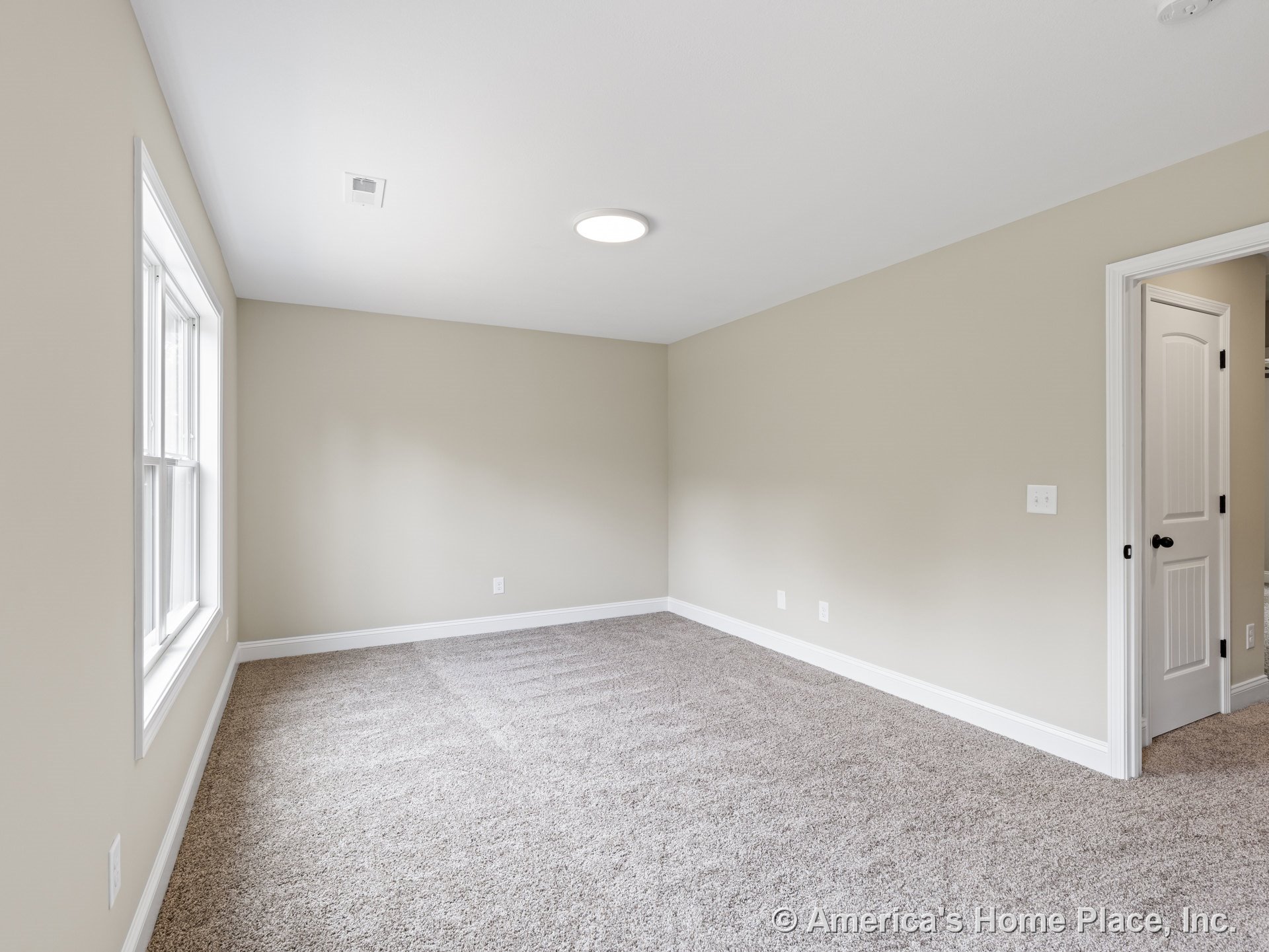 Upstairs bedroom with beige carpet flooring, light tan walls, large window with white trim, flush ceiling light, and paneled door.