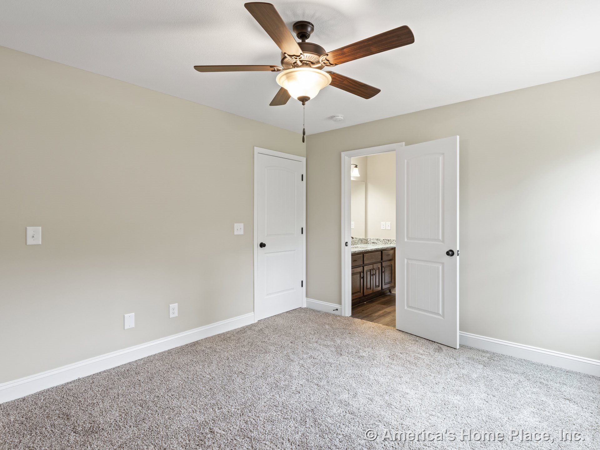 Beige carpeted bedroom with ceiling fan and light fixture, white paneled doors, neutral wall color, and en suite bathroom featuring granite countertop and dark wood cabinetry.