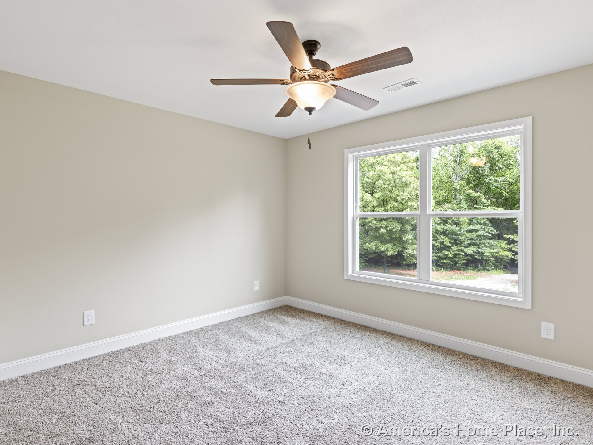 Beige carpet flooring, ceiling fan with integrated light fixture, large double window allowing natural light, neutral wall color, white baseboard trim, rectangular room layout.