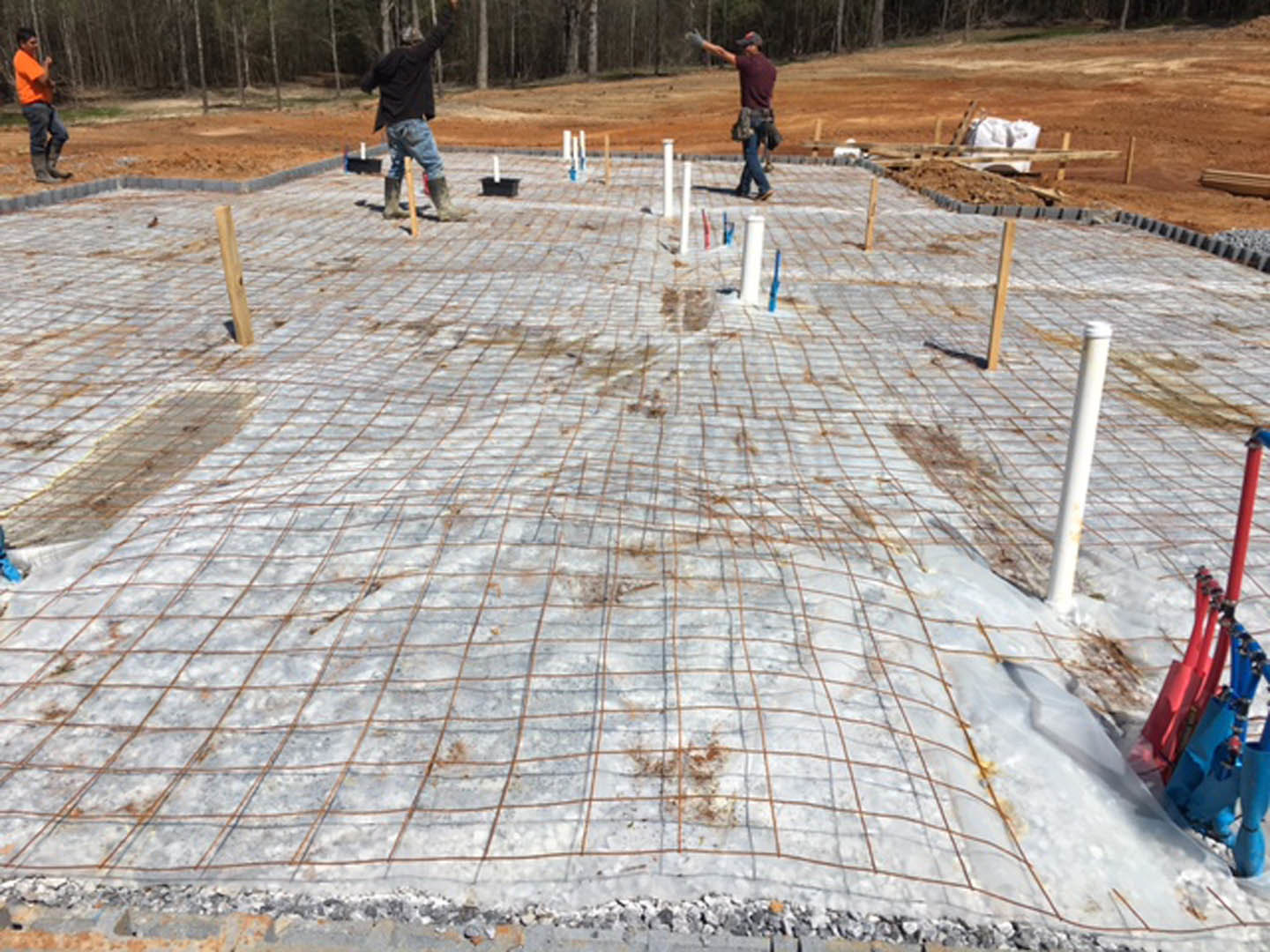 Framed exterior walls and foundation under construction, three men in orange, black, and red shirts working on site, surrounded by trees and building materials