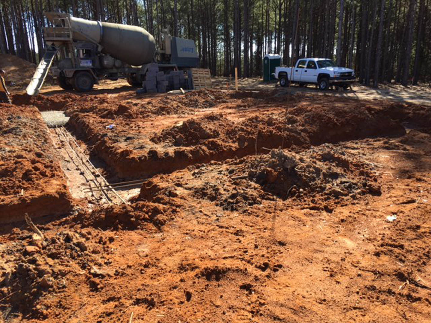 White truck and cement mixer parked on dirt construction site with muddy ground, scattered trees, and tire tracks