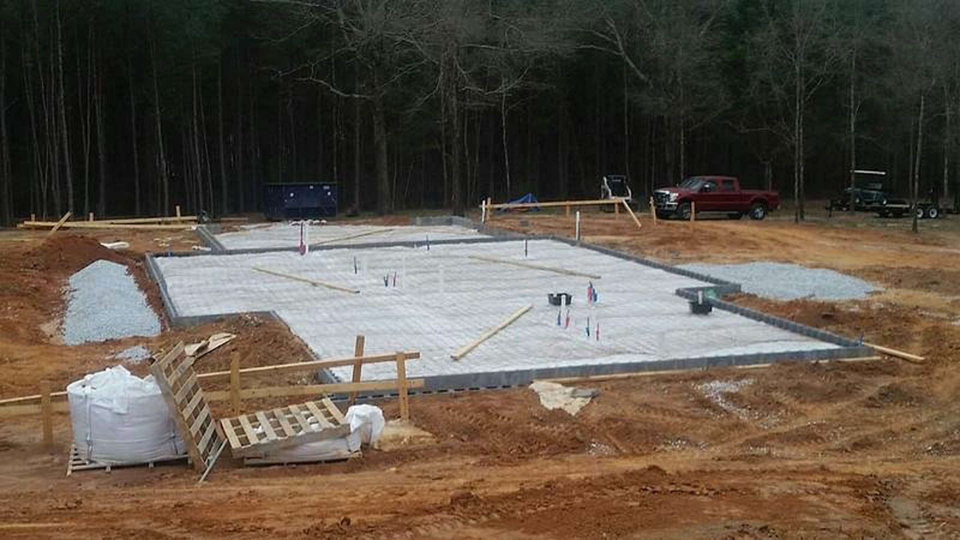 Concrete foundation slab at a residential construction site with a red car, truck, pallet, large white bag, and scattered building materials; trees in the background.
