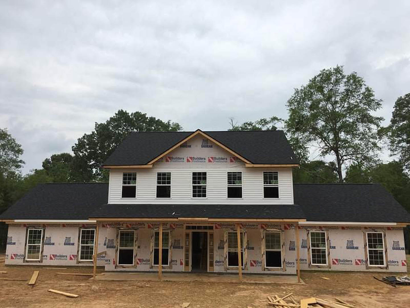 Partially built house with black shingle roof, multi-pane windows, and black glass door, surrounded by trees under cloudy sky