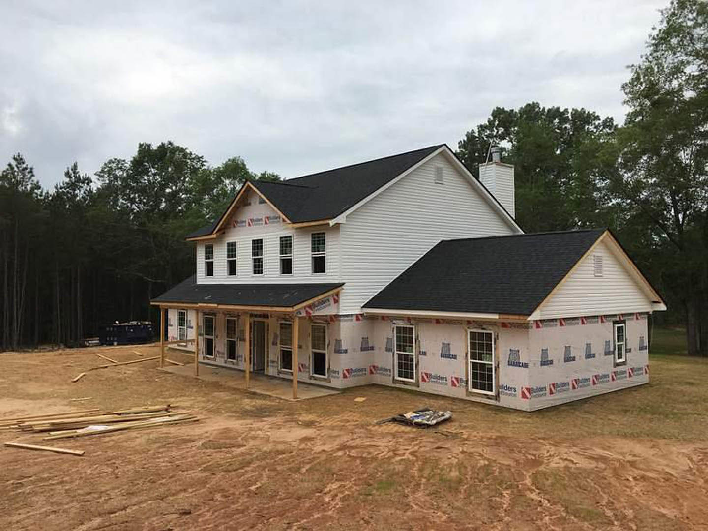Partially built house with black roof and white-framed windows, surrounded by trees, construction materials and wood pile scattered on bare ground
