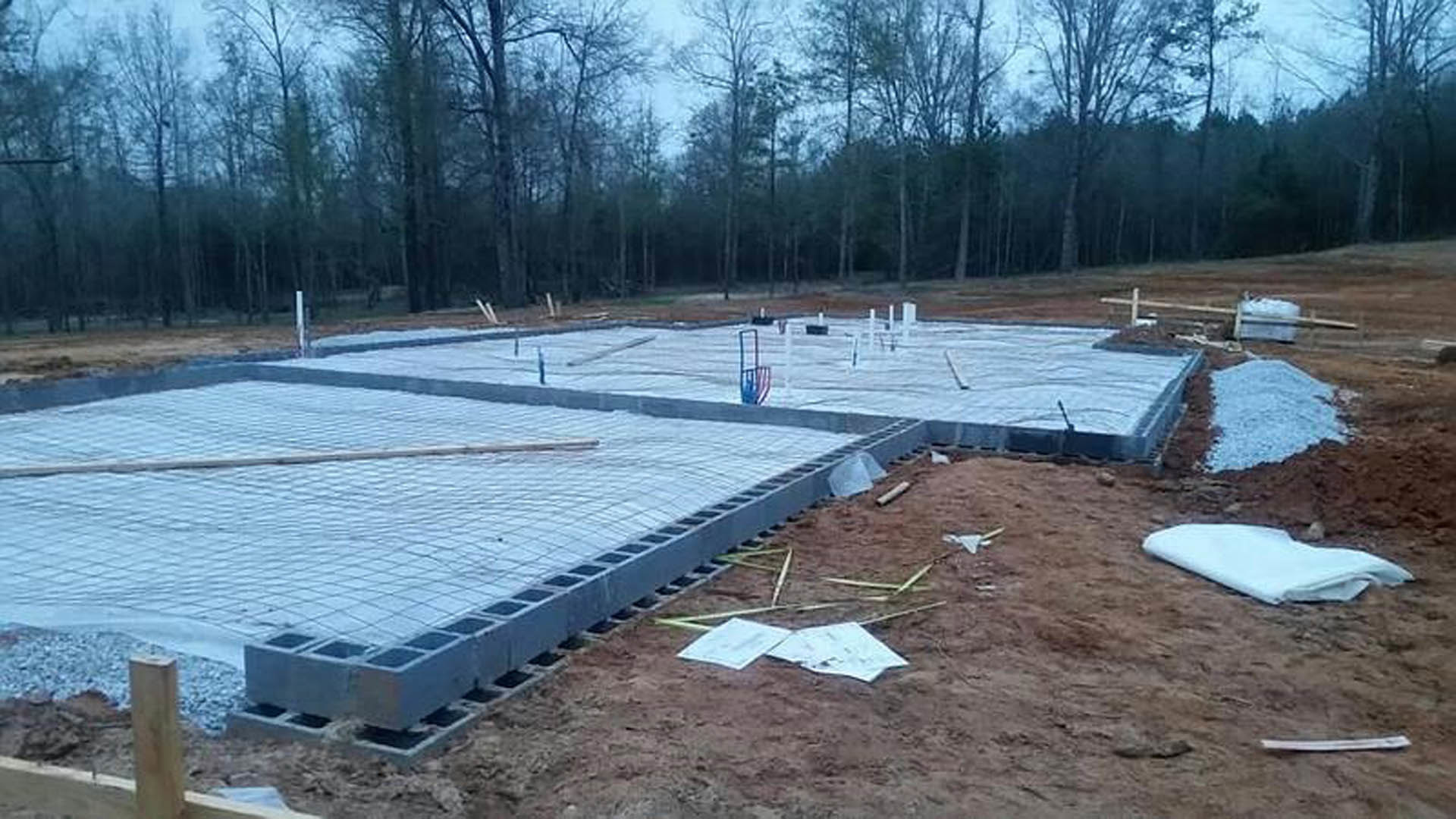 Concrete foundation under construction with grid pattern, white protective sheeting covering ground, surrounded by trees and patches of snow
