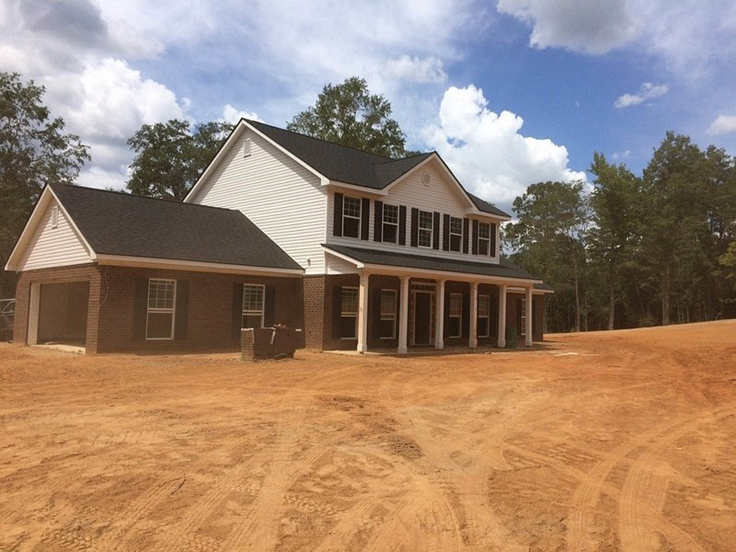 Single-story house with white roof, large windows, and dirt yard; mature trees and cloudy sky in background