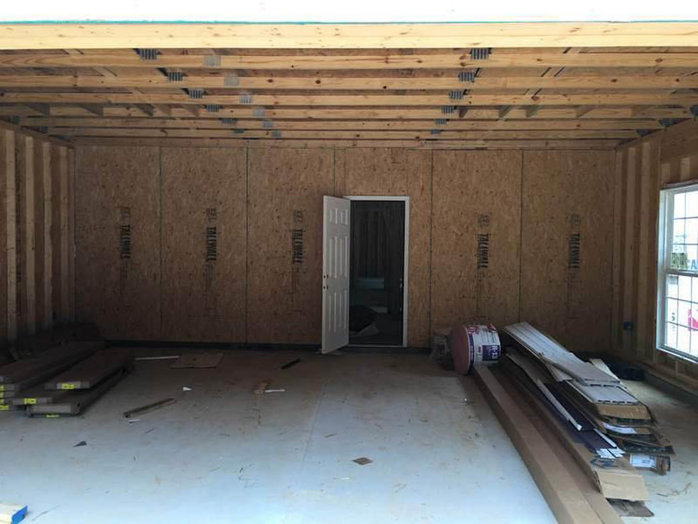 Open white door with black knobs, dark curtain partially visible, pile of lumber stacked on light wood floor, unfinished wood wall, plaster ceiling, and exposed beams in