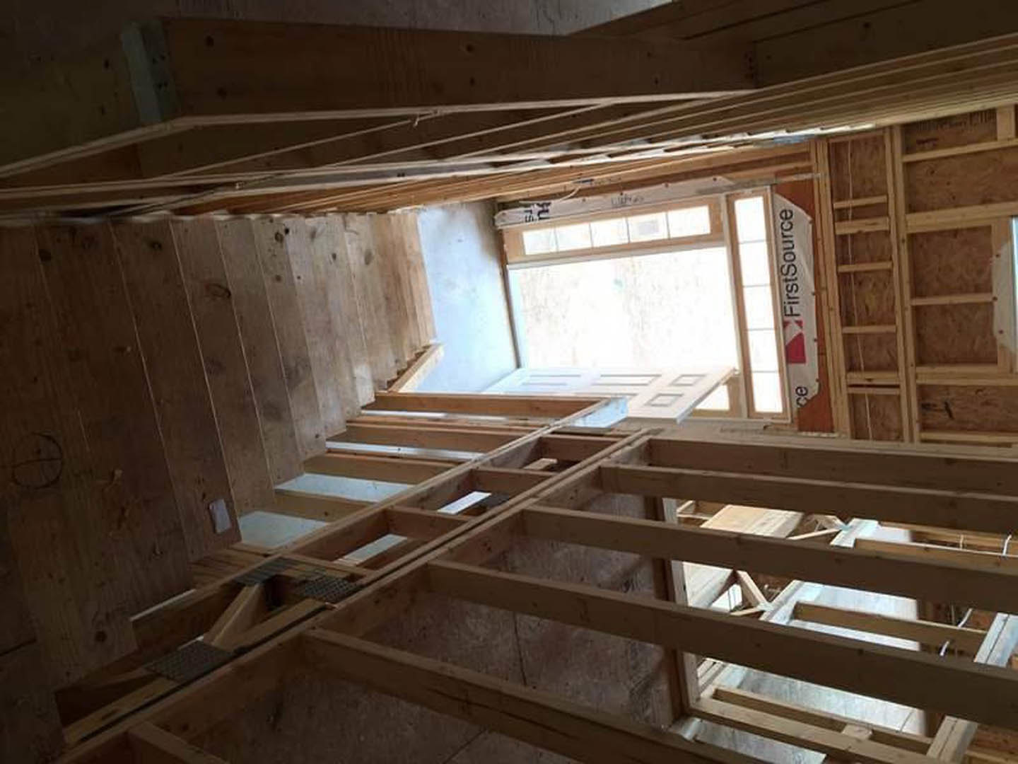 Exposed wood framing with plywood walls, wooden ladder leaning against studs, window opening and door frame visible, white construction sign with black text attached to framing