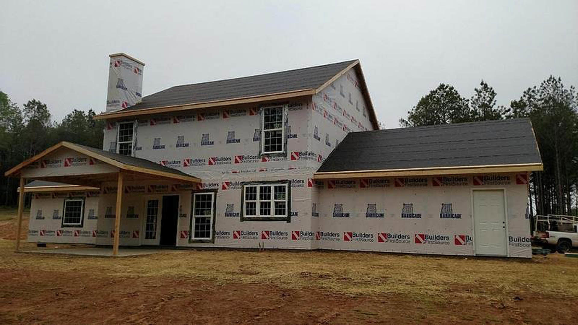 Partially built house with exposed plywood walls, black-trimmed white door, several installed windows, shingled roof, dirt yard, and white pickup truck parked near trees in