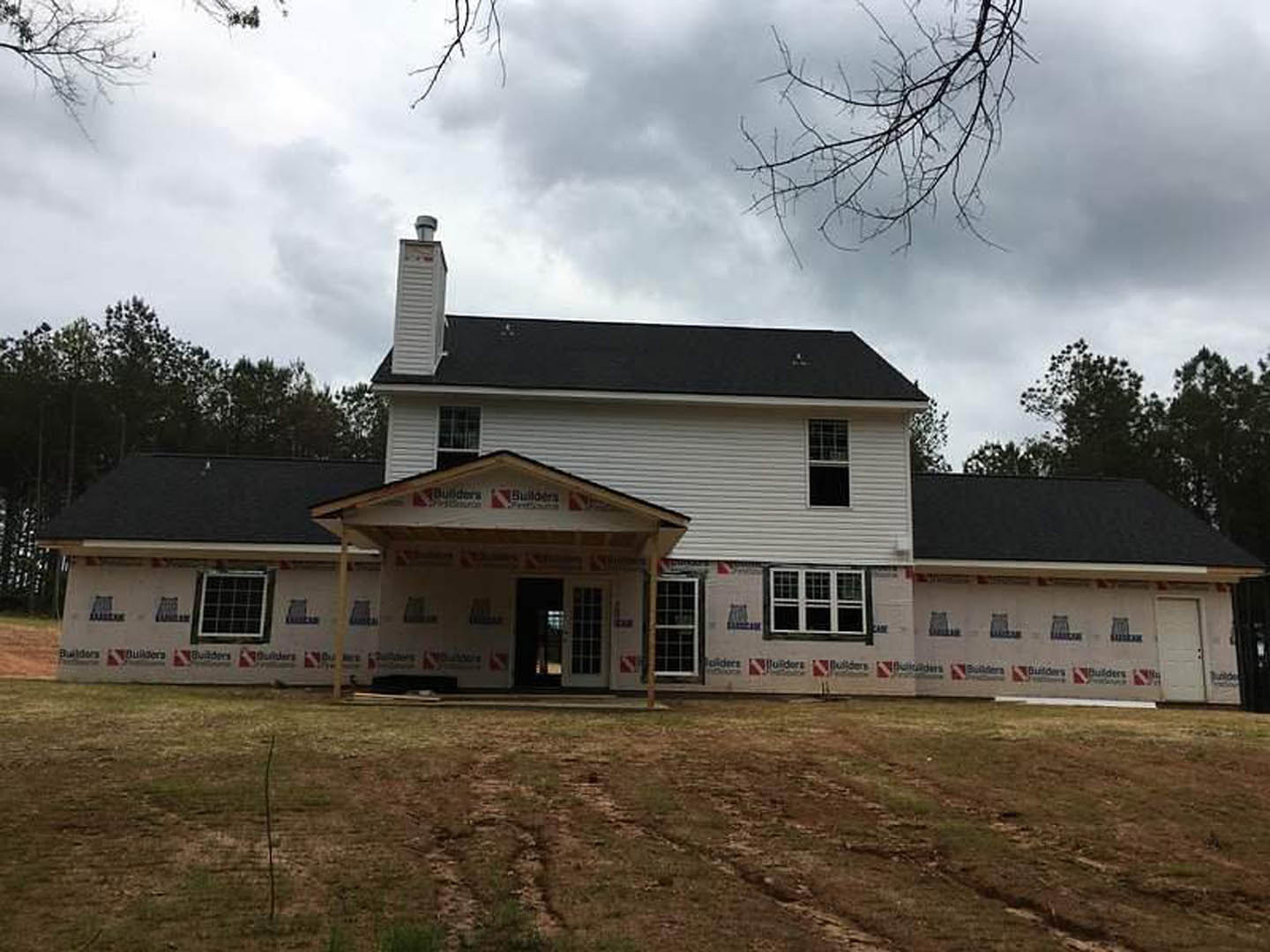 White farmhouse-style home with covered front porch, square grid windows, exposed wood beams, and dirt yard under cloudy sky
