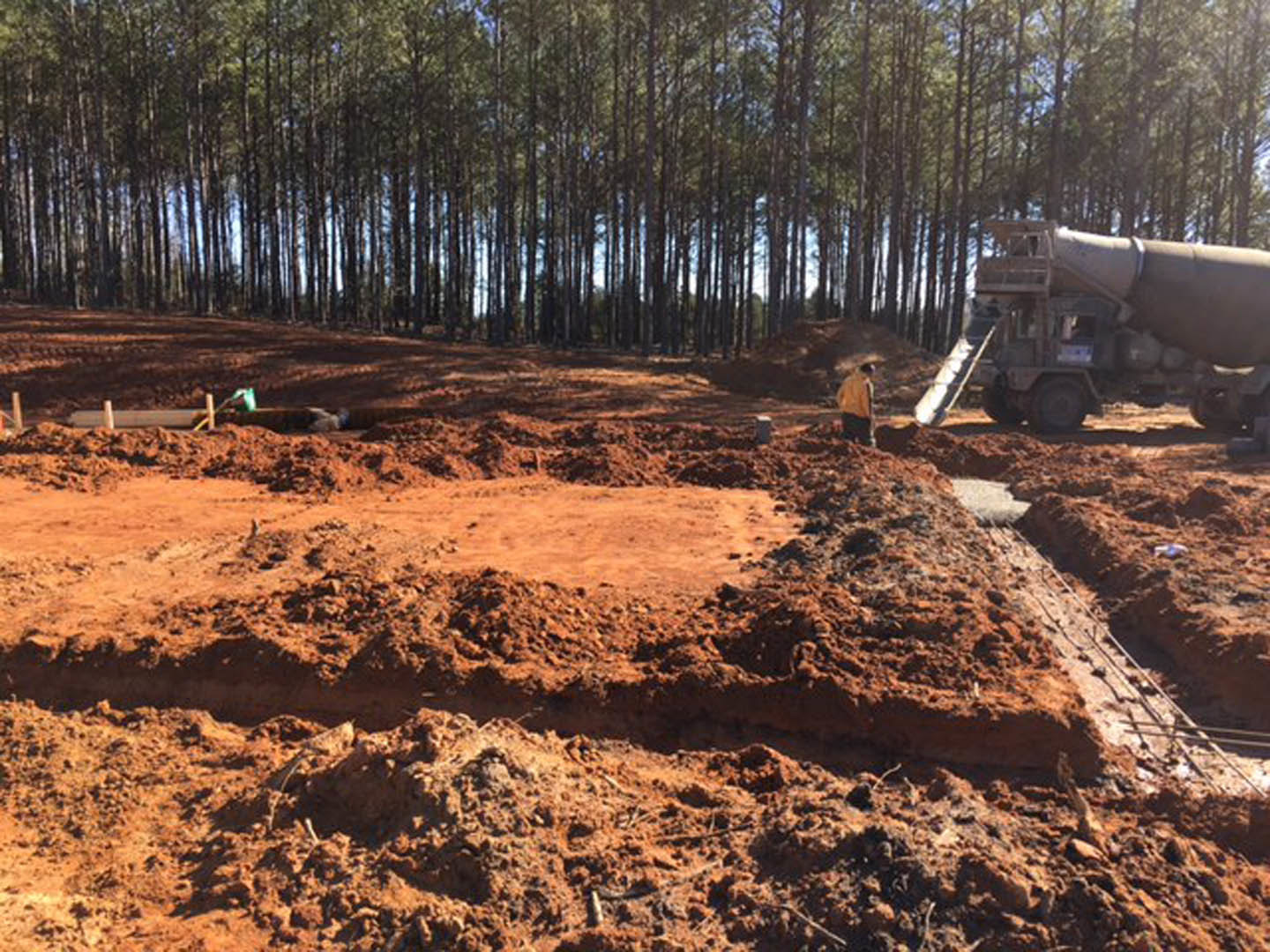 Construction site with cement mixer truck parked on dirt ground, man standing near soil pile, surrounded by tall trees and muddy terrain