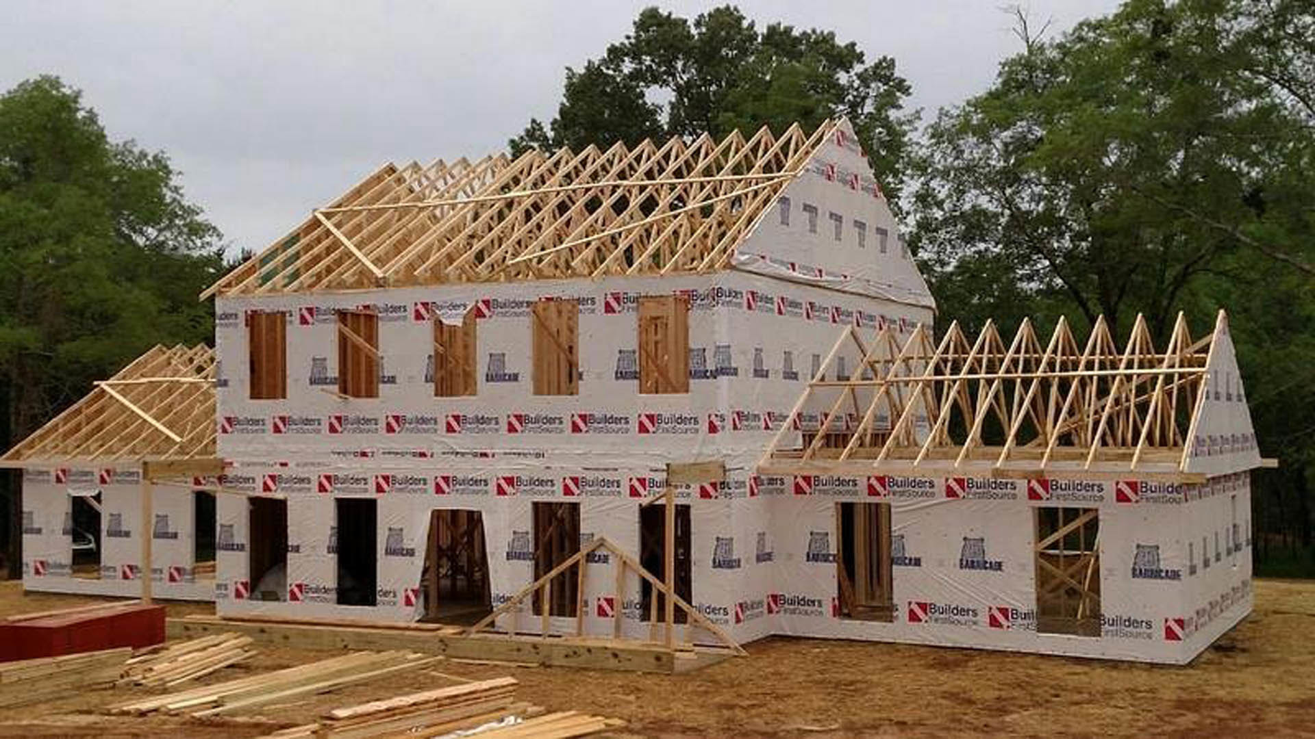 Wood-framed house under construction with exposed beams, partially completed exterior walls, and open window spaces, set outdoors with trees and sky in the background