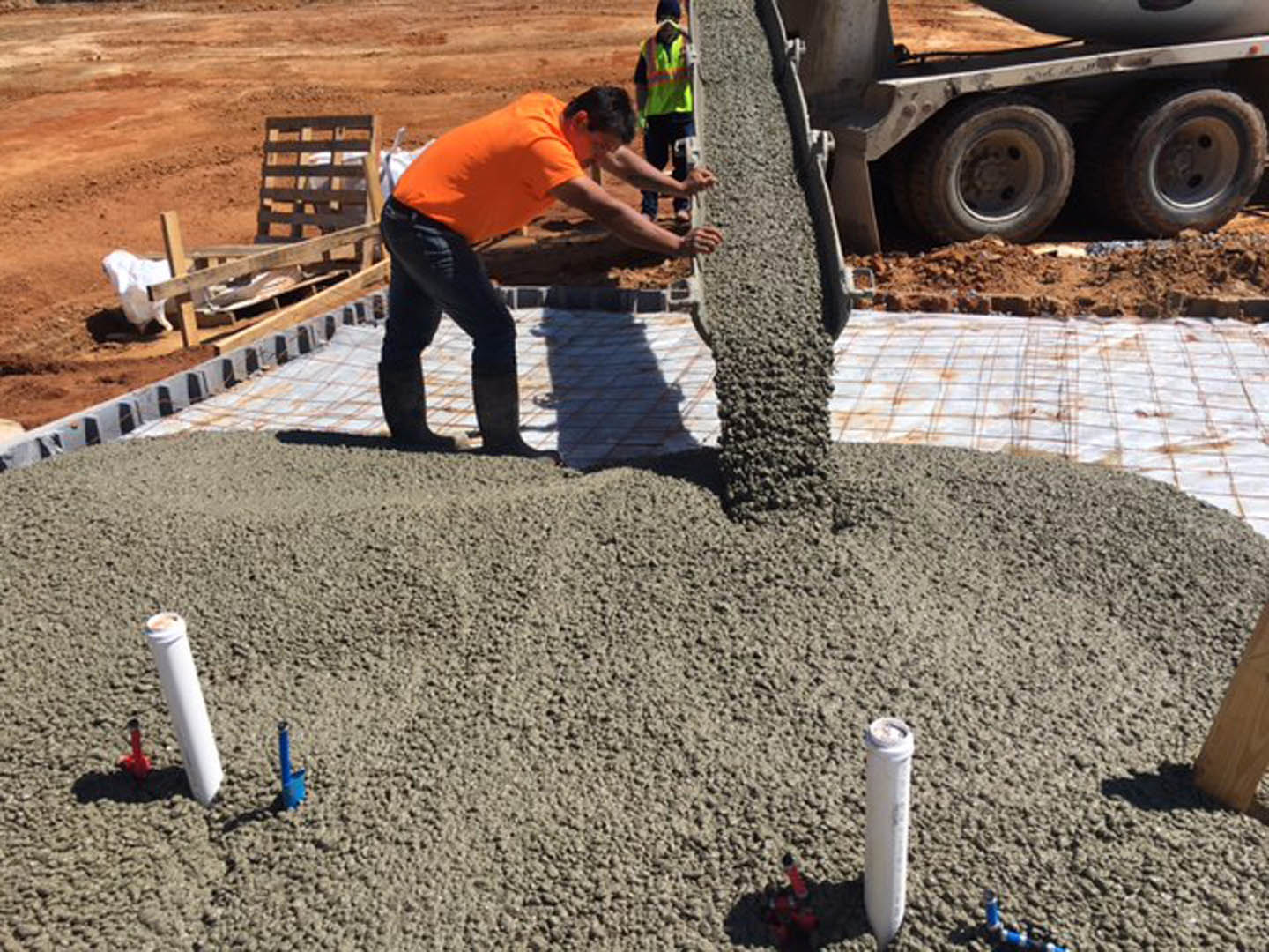 Man in orange shirt pouring cement onto gravel next to wooden pallet and wheel on dirt construction site