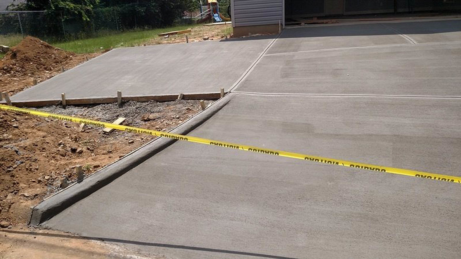 Yellow caution tape stretched across concrete driveway bordered by asphalt road, curb, and trees.