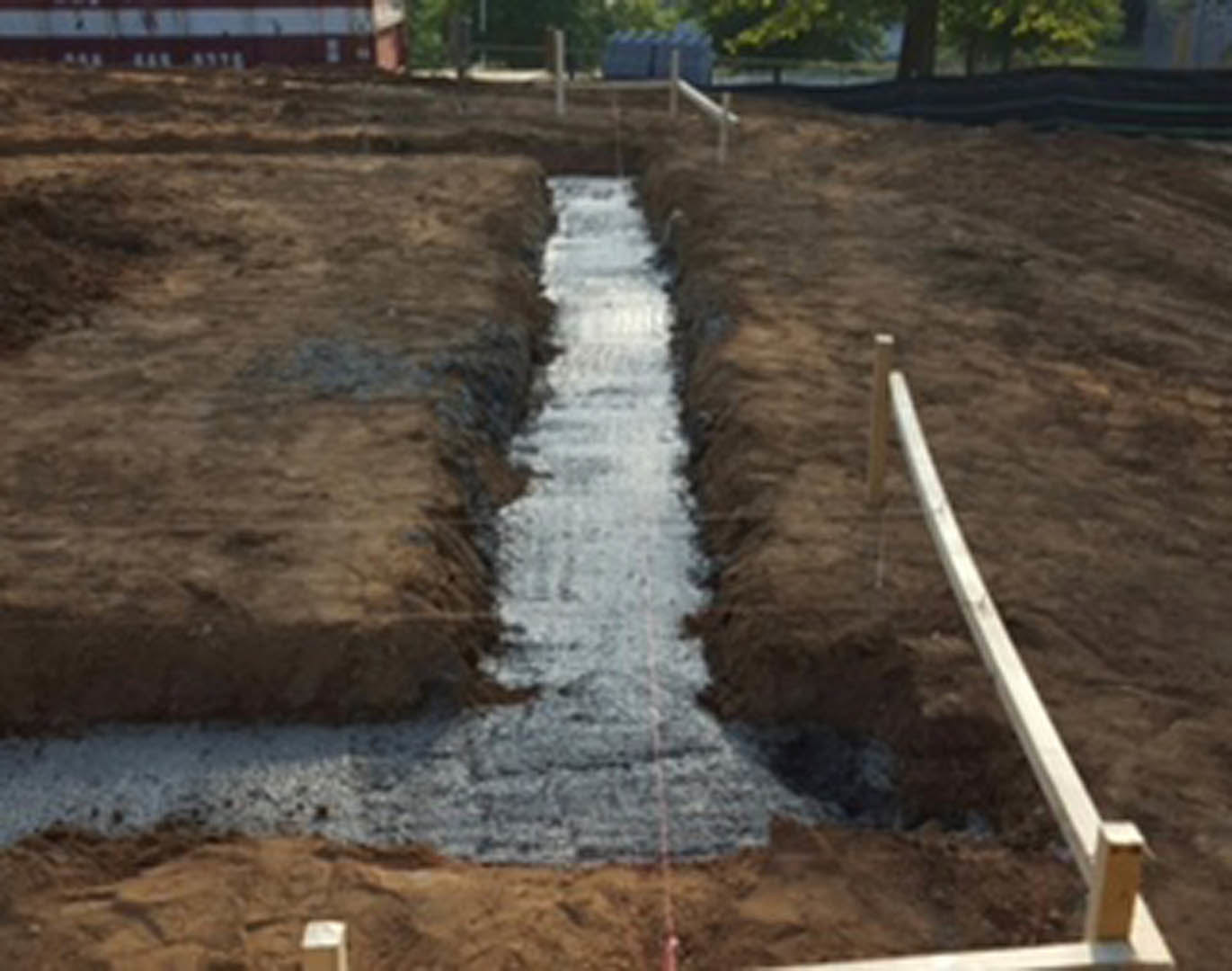 Deep trench dug in brown soil beside a wooden fence, with blurred building and tree trunk in the background