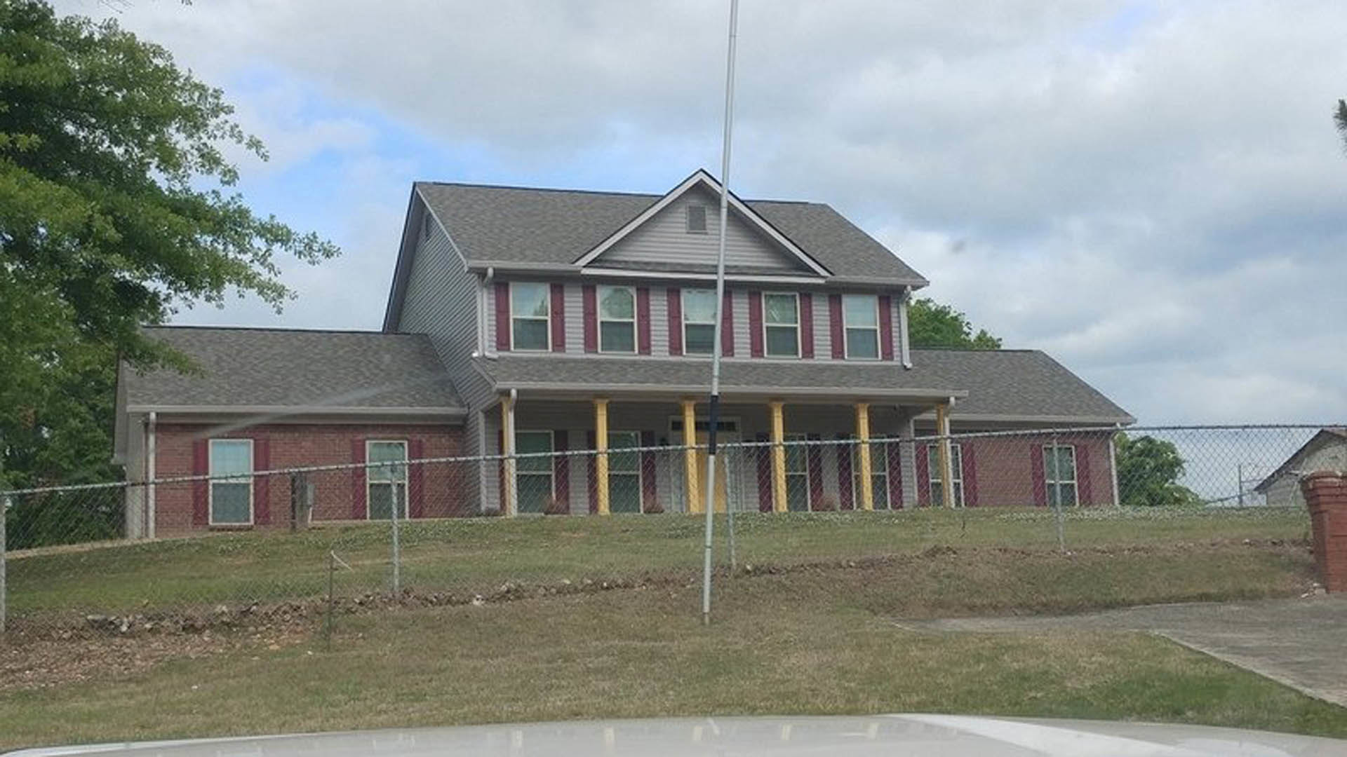 Two-story brick house with white-framed windows, wooden porch, fenced yard, green grass, and mature tree under a blue sky with scattered clouds