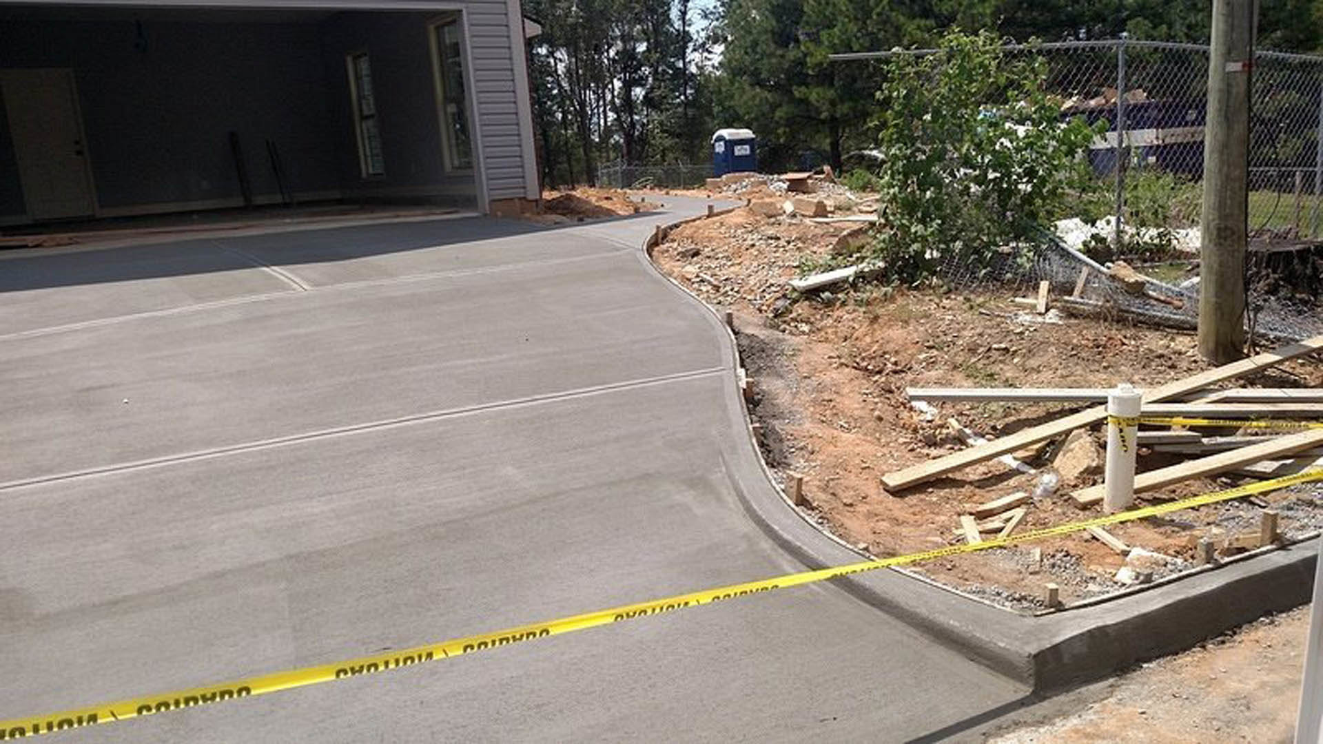 Yellow caution tape stretched across concrete driveway in front of house, with tree and wood debris visible near fence.
