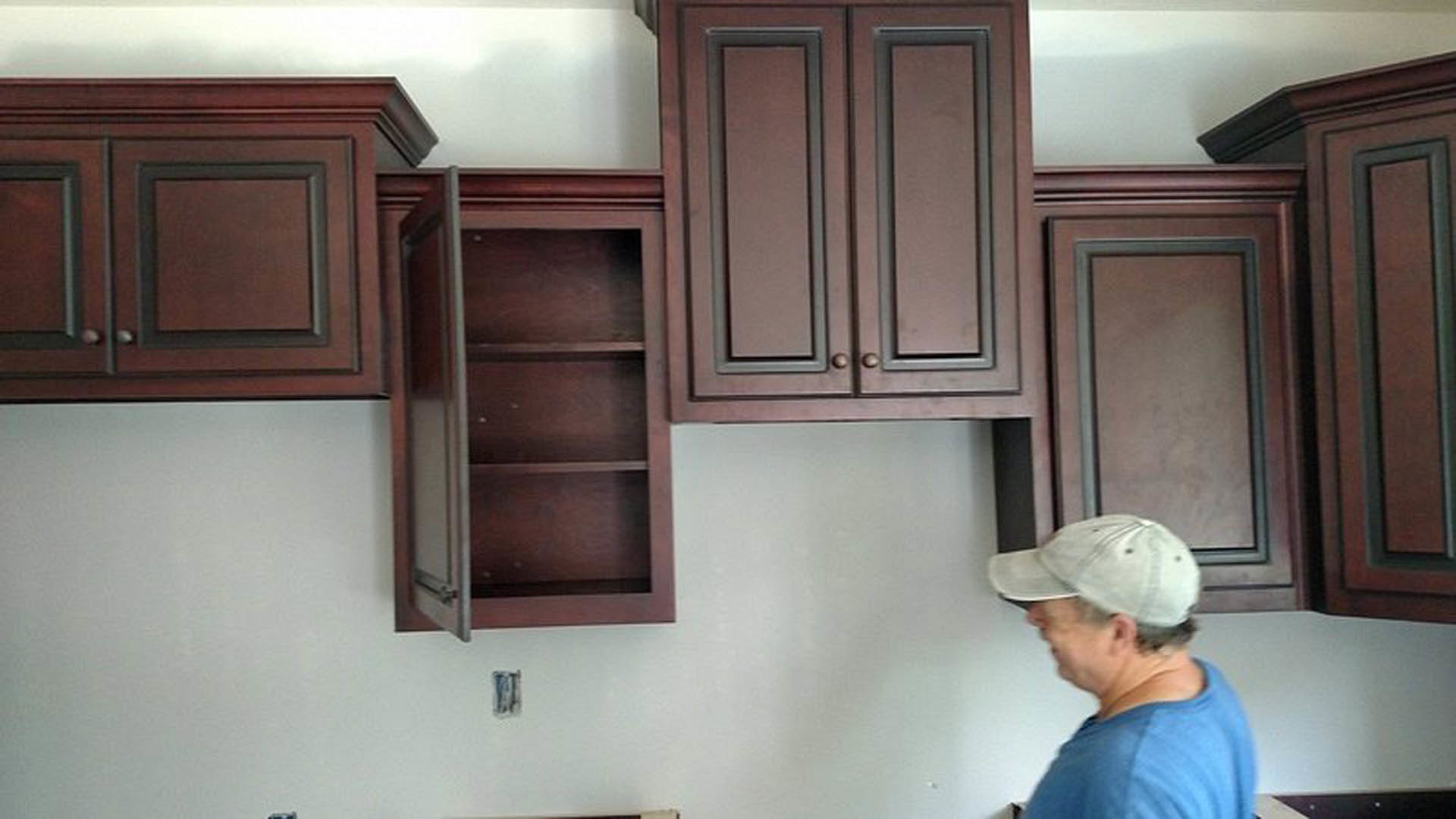 Man in blue shirt and baseball cap standing in front of unfinished kitchen wall with empty wooden cabinets and open shelving