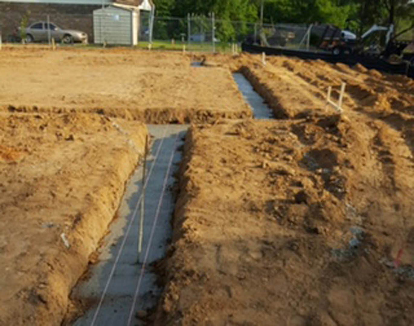 Foundation stage of custom home construction with several square holes in muddy soil, surrounded by scattered concrete slabs, trees, and construction vehicles.