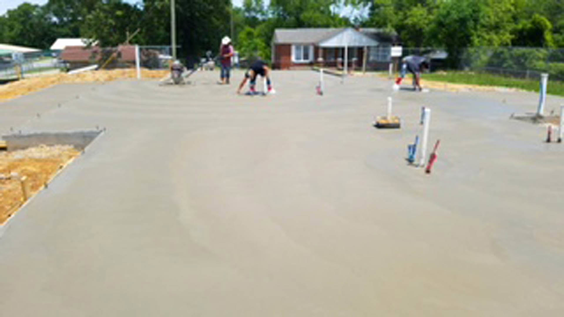 Workers pouring and smoothing concrete slab foundation outdoors, surrounded by trees and open sky