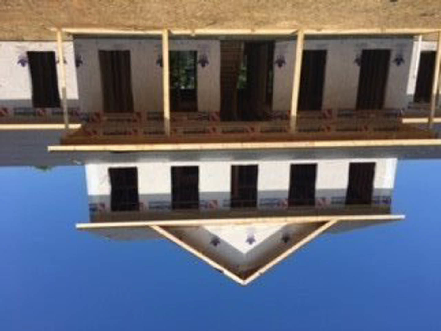 Wood-framed custom home under construction with exposed beams, unfinished exterior walls, and large windows reflecting blue sky