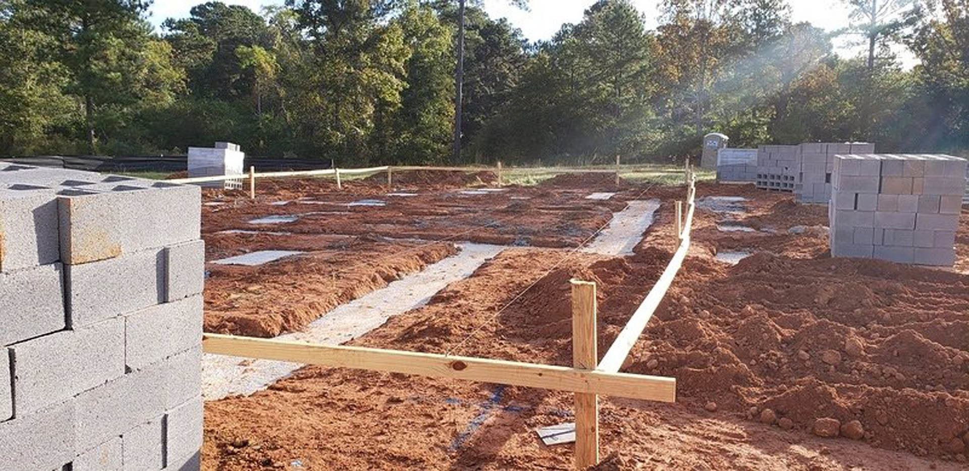 Dirt-filled construction site with wooden foundation posts, scattered soil, and surrounding trees under a clear sky