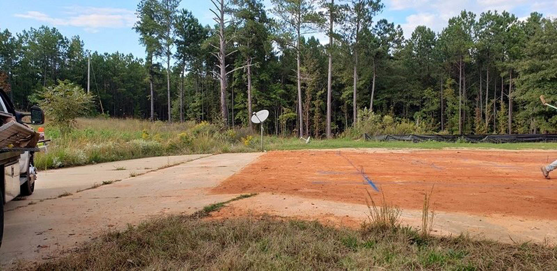 Modern custom home with standing seam metal roof, surrounded by grassy field and scattered trees, satellite dish mounted near exterior, dirt driveway visible in foreground