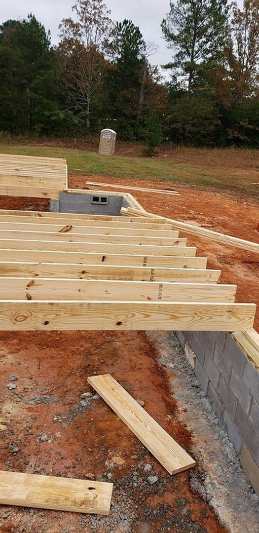 Exposed wooden beams under construction on a residential site, with lumber planks and foundation visible against an outdoor backdrop