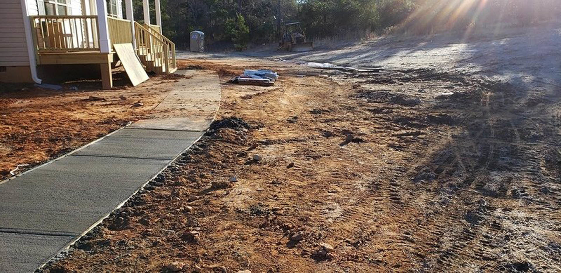 Dirt road with tire track leading to a wooden porch with ramp, surrounded by trees and plants, wooden board attached to exterior structure