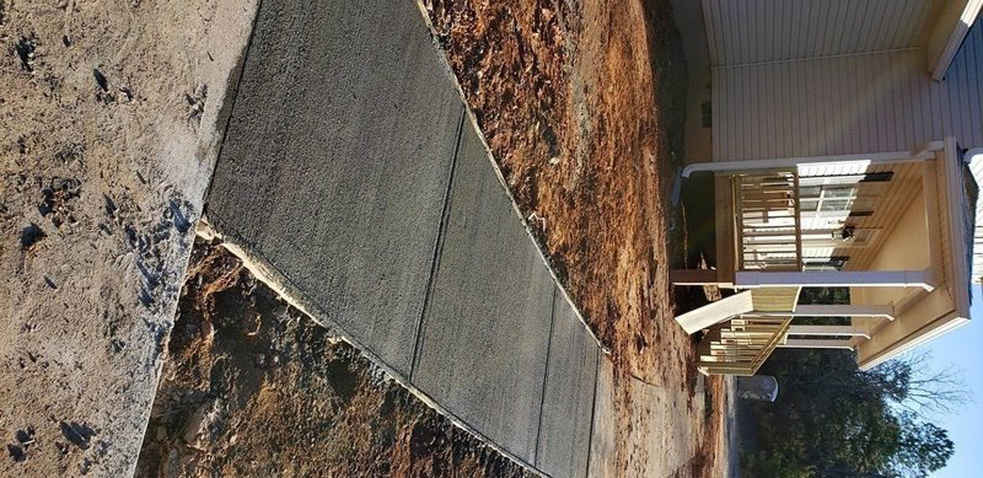 Two-story house with white siding and large windows, wooden porch with white railing, wooden structure beside the road, mature trees in the yard, person walking on sidewalk holding