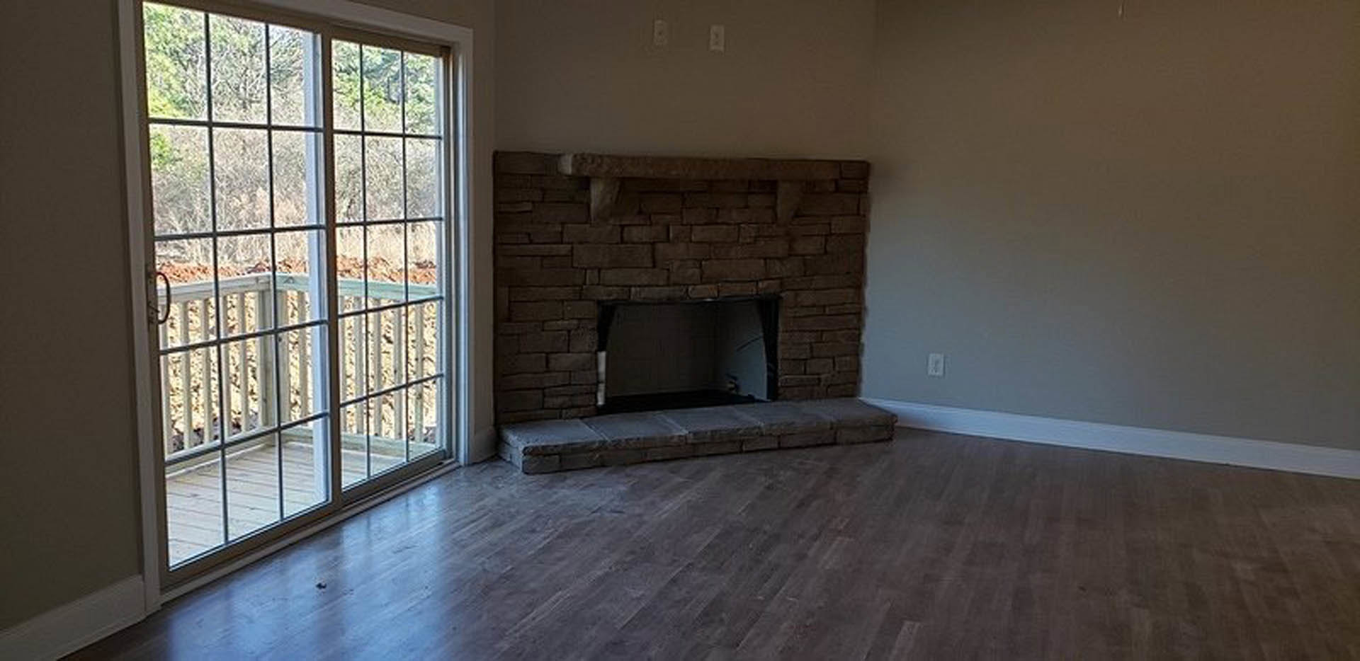 Brick fireplace with stone hearth, wooden flooring, sliding glass door opening to balcony, partial view of kitchen with person standing