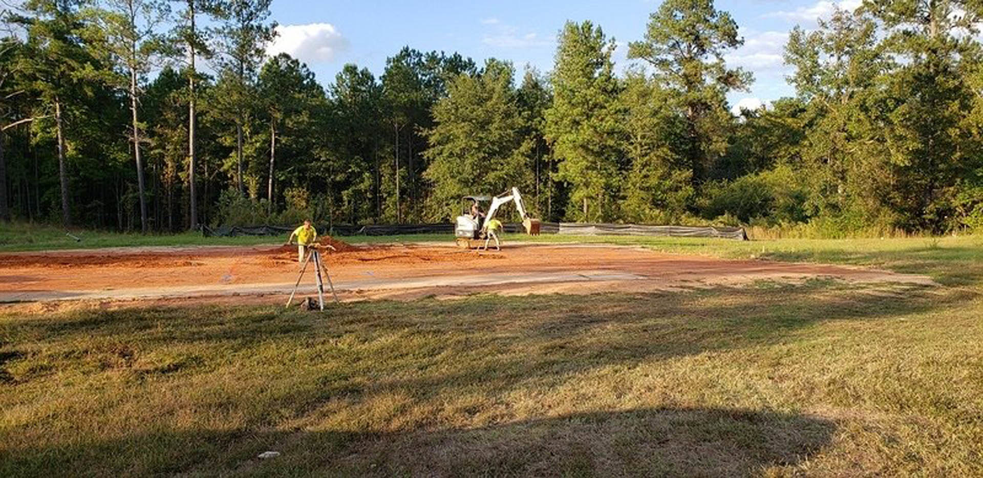 Construction site with a yellow tractor on dirt, man in yellow shirt operating surveying tripod, grassy area and trees in background