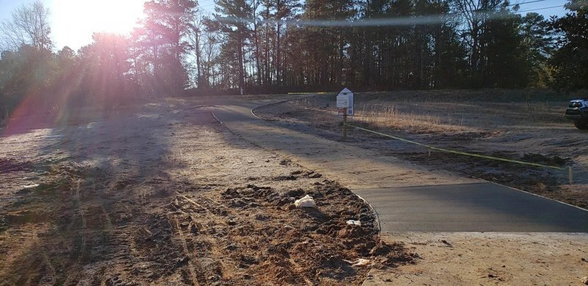 Dirt road bordered by trees and plants under morning sunlight, with a blurry car in the background