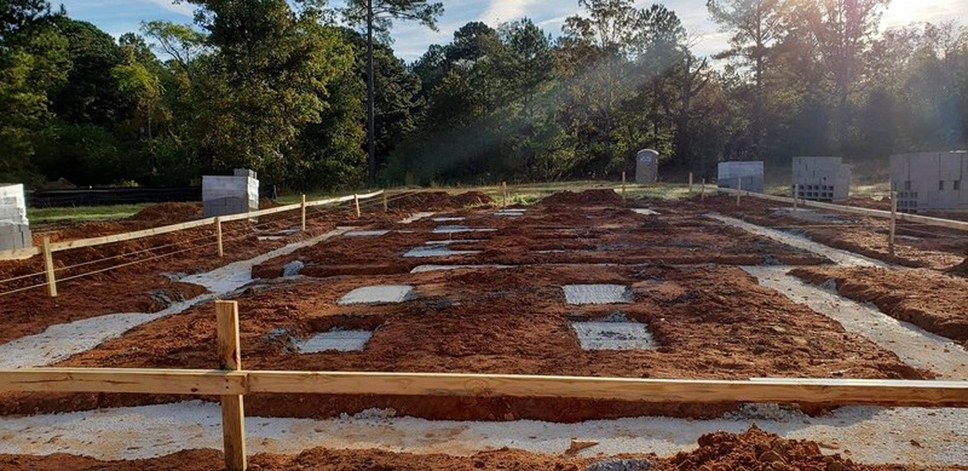 Dirt-covered construction site with scattered square tiles, wooden board lying on soil, trees and plants in background under open sky
