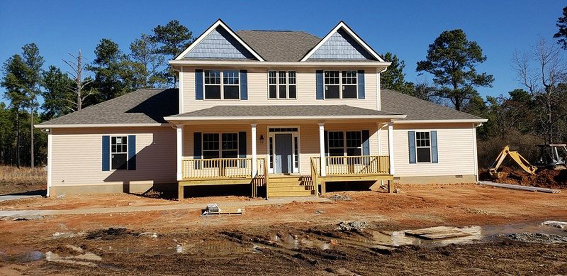 Wooden deck and porch railing on a partially constructed house, grid windows and door visible, dirt patch in front yard, siding and cottage-style exterior details.
