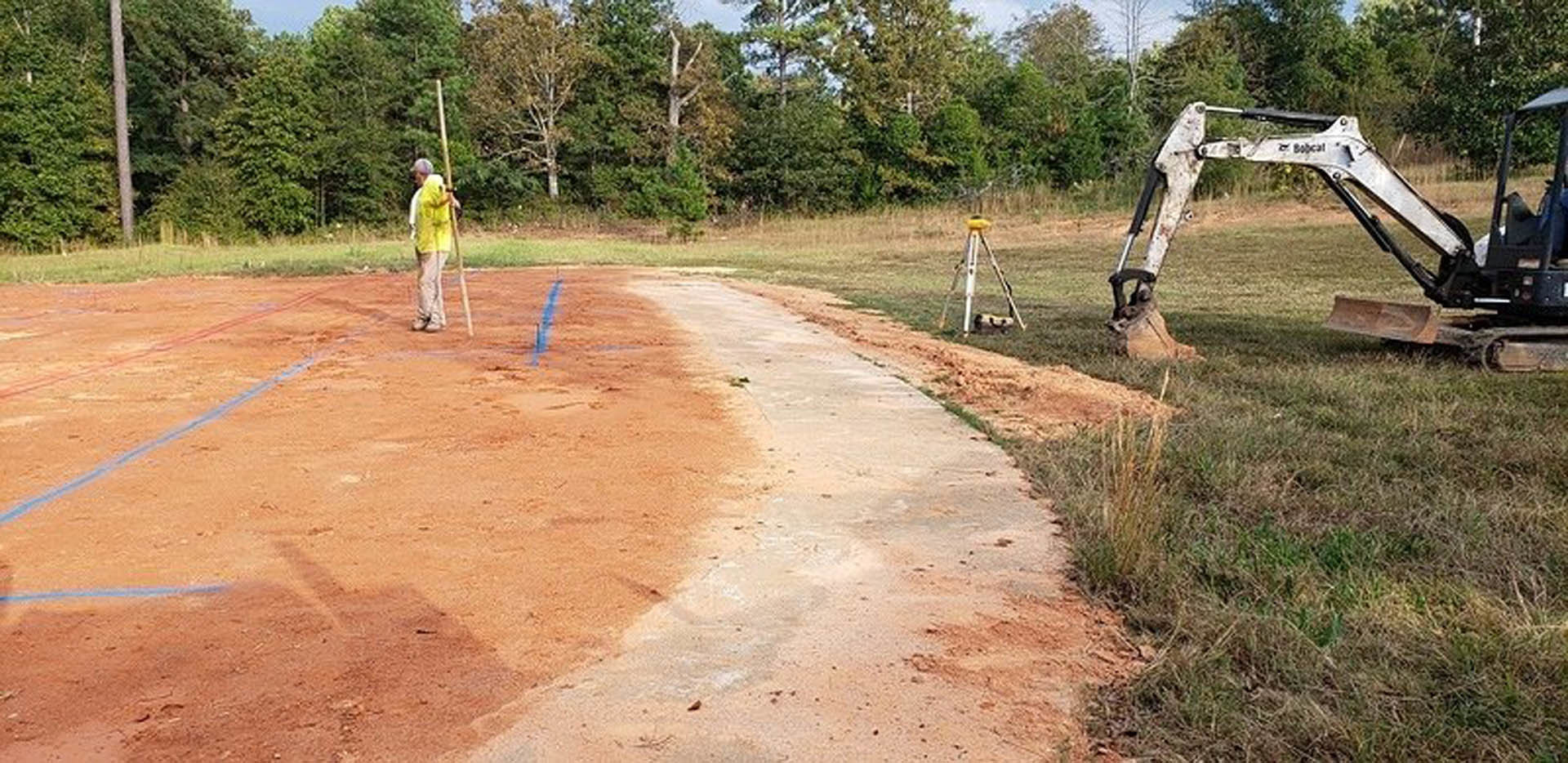 Man standing beside a dirt path near construction equipment, surrounded by grass, trees, and soil under a partly cloudy sky