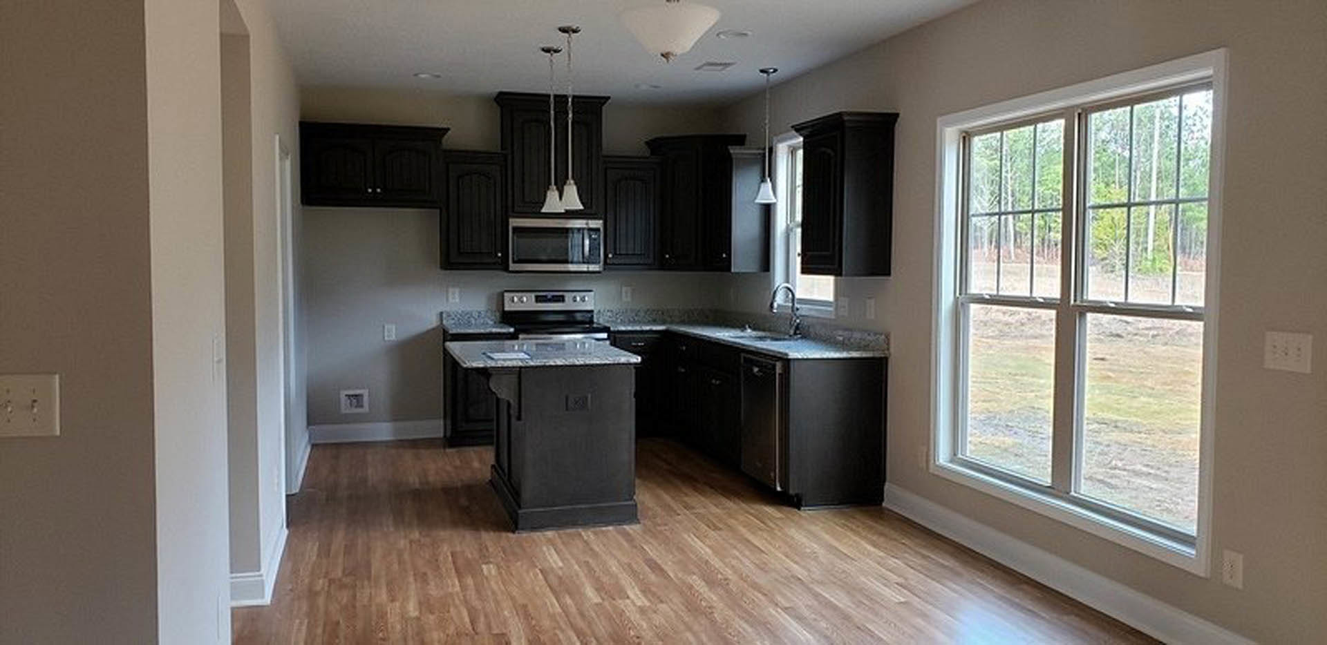Black kitchen cabinets with brushed metal hardware, wood plank flooring, white marbled countertop, stainless steel sink, and large window providing natural light.