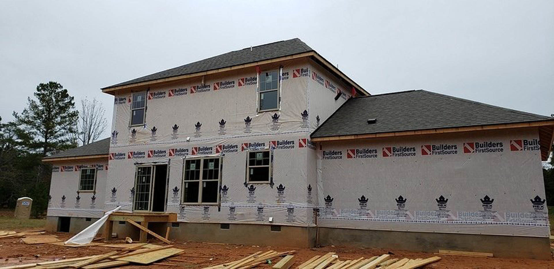 Partially built house with exposed wooden framing, several installed square-paned windows, and construction materials scattered outside