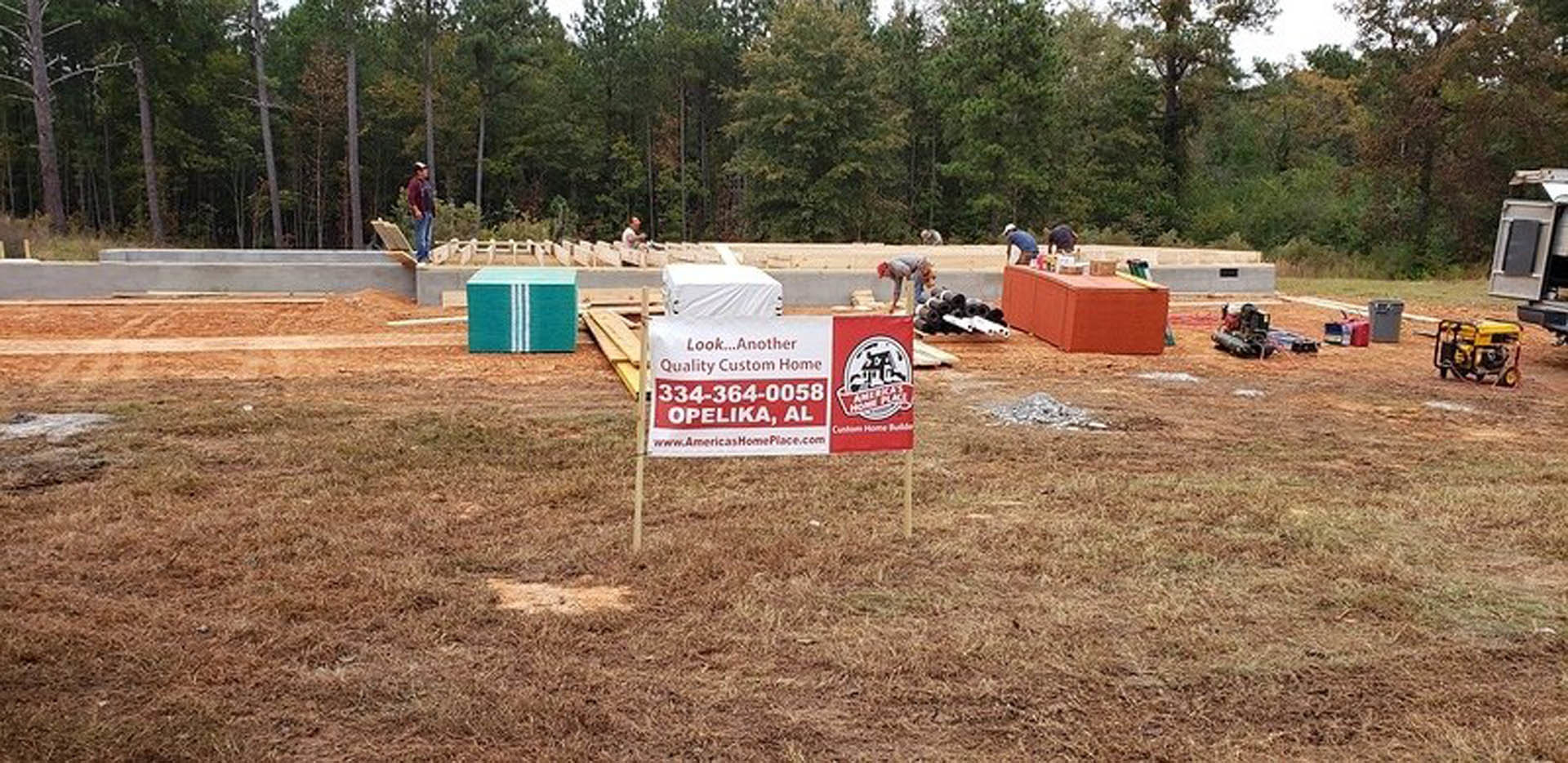 Construction site with exposed soil, grass, and trees; workers near a black rectangular object on gray pavement; yellow construction vehicle and a sign visible in the foreground