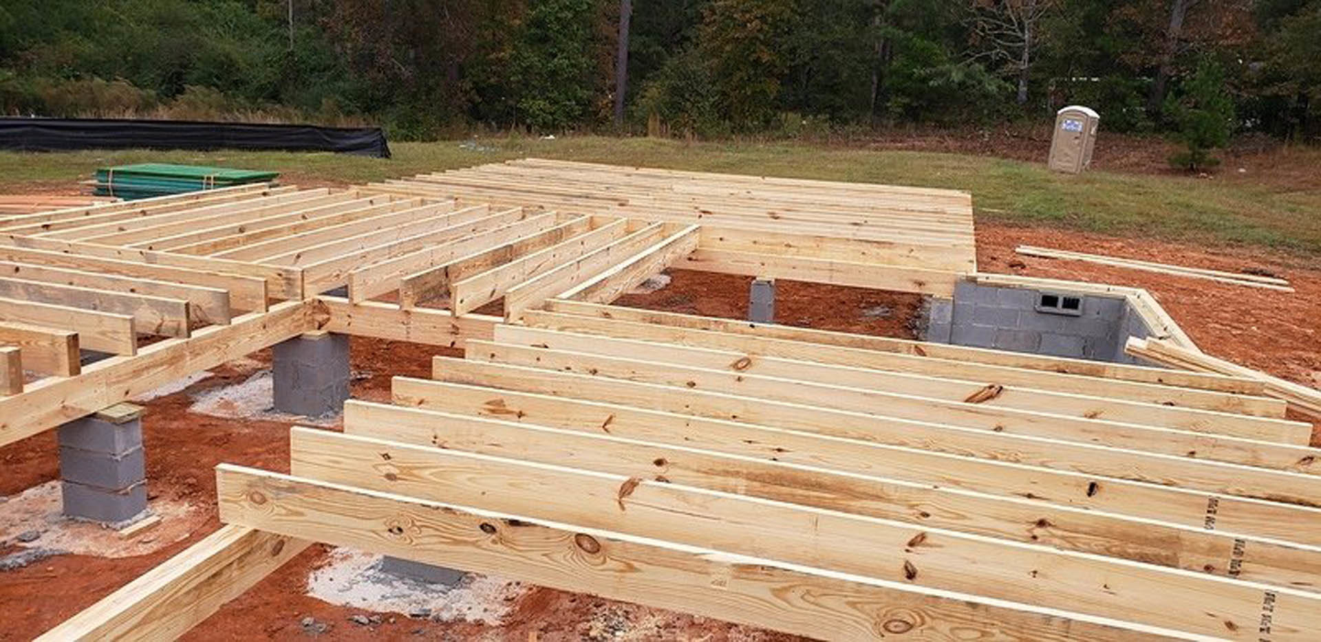 Wood-framed house under construction with exposed lumber, partially built wooden steps, portable toilet, and scattered building materials; green bench and car visible in