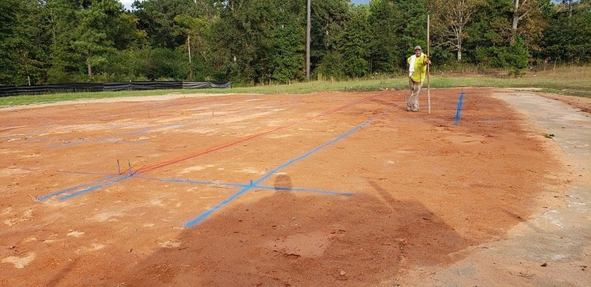 Man wearing yellow shorts standing in a dirt field with trees and playground equipment in the background under a clear sky