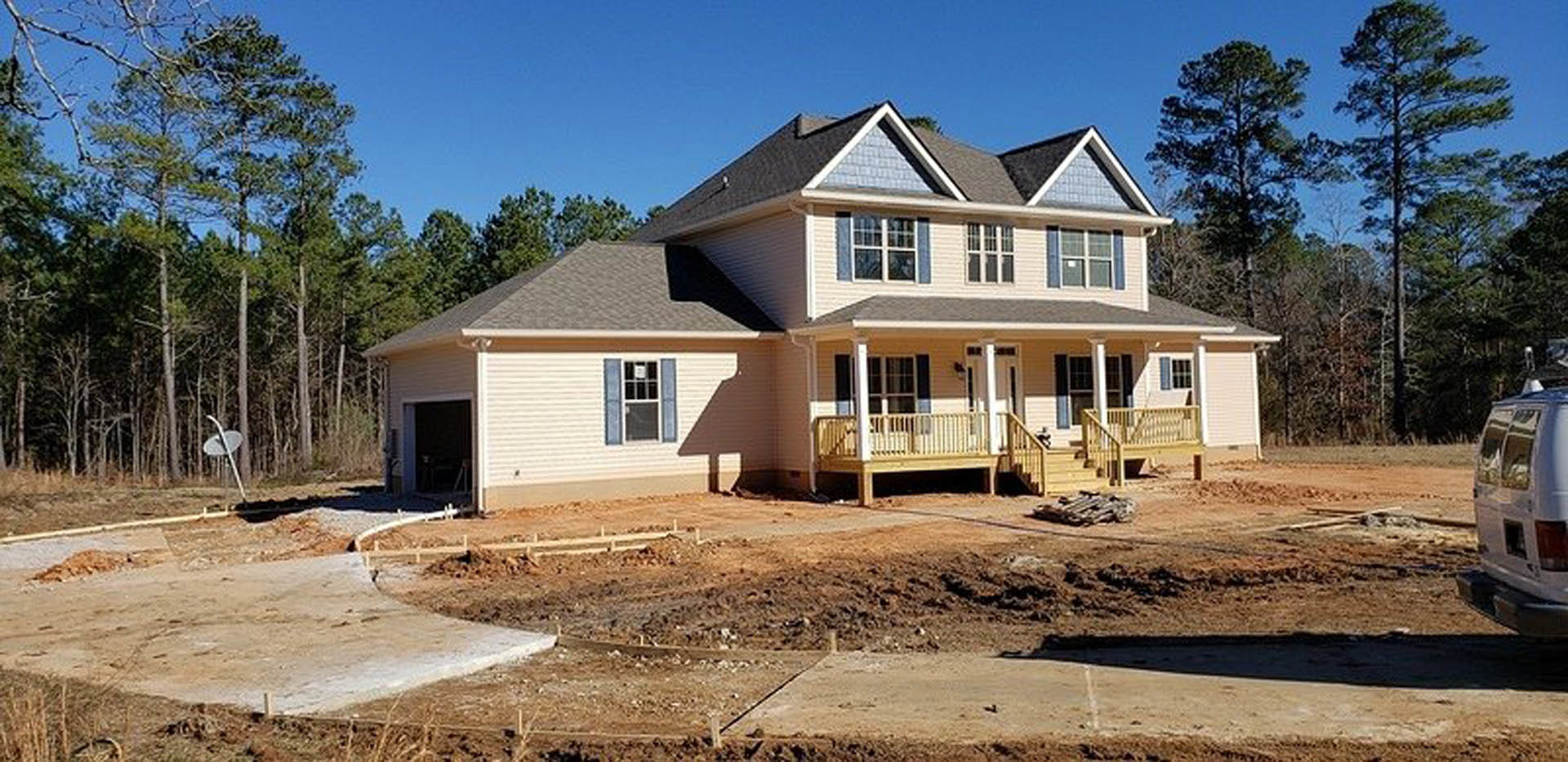 Two-story custom home under construction with exposed dirt yard, white van parked in front, unfinished porch with metal railing, multi-pane windows including one with a