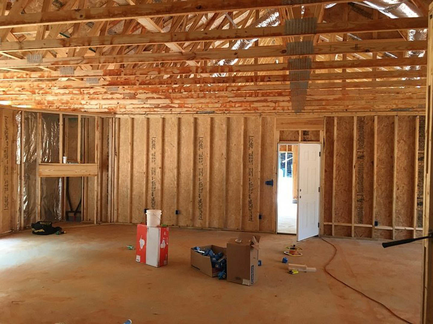 Exposed wooden beam ceiling, white door with bright light, unfinished walls, construction materials including a red box with toilet paper and a white bucket on the floor