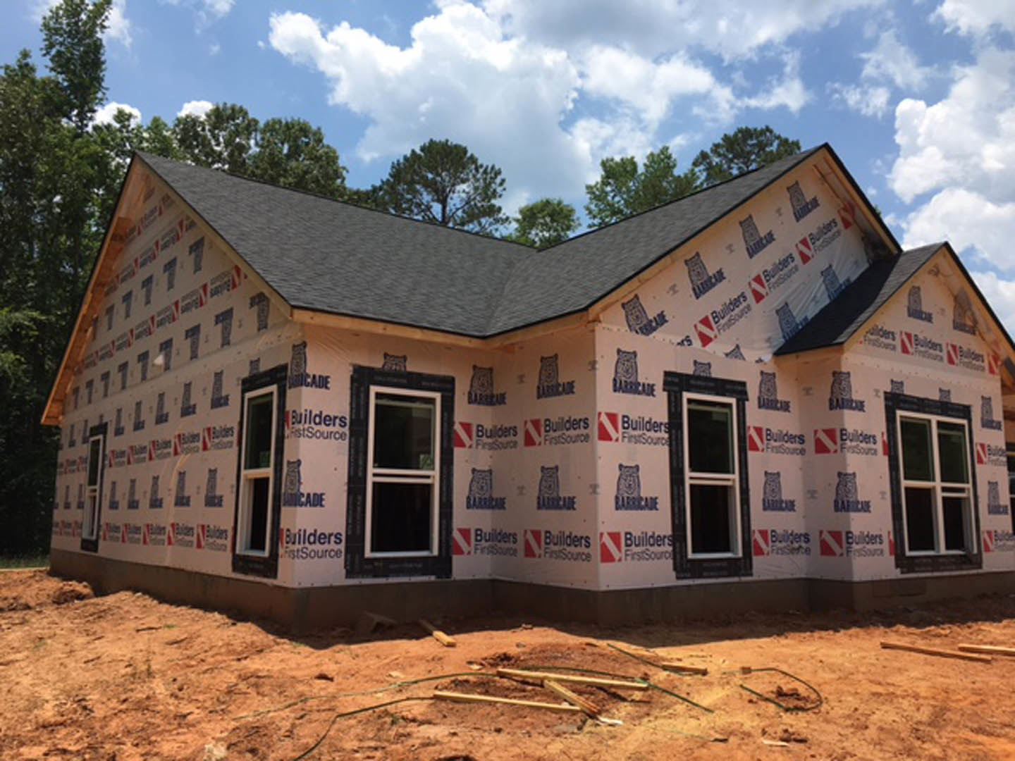 Partially built house with exposed insulation, white-framed windows, scattered wood pieces on ground, surrounded by trees under a blue sky with clouds