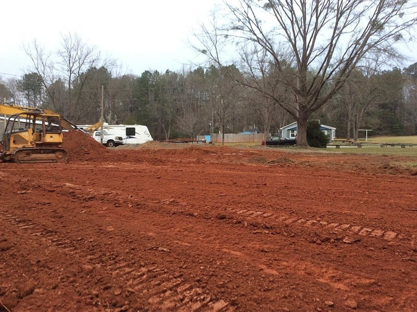 Construction site with exposed soil, tractor parked on dirt field, tire tracks visible, large tree and tree trunk near a trailer, white RV in background, open sky overhead