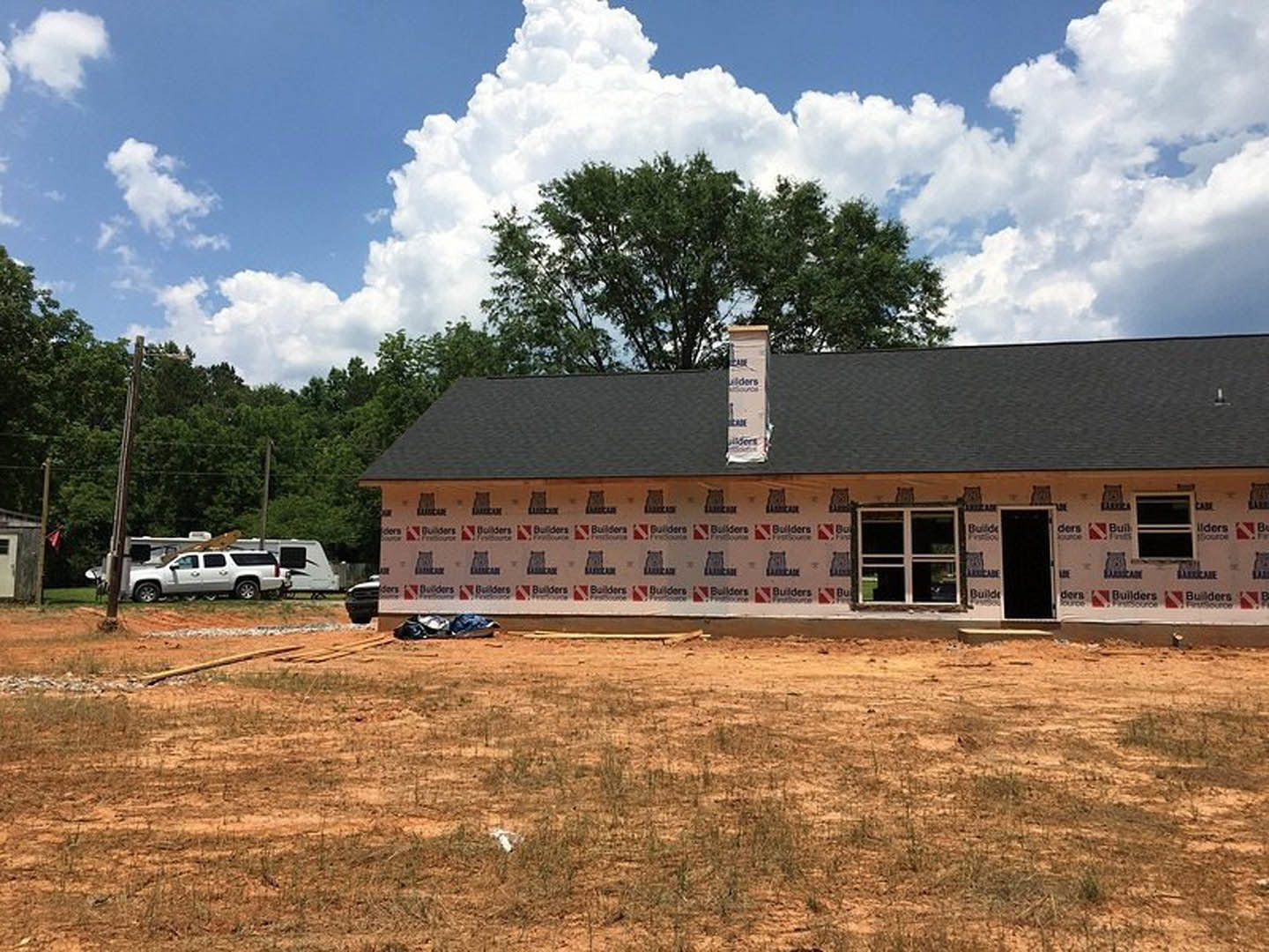 Partially built house with exposed framing and chimney, surrounded by dirt, grass, and mature trees under a blue sky with scattered clouds; white truck parked nearby, red and white